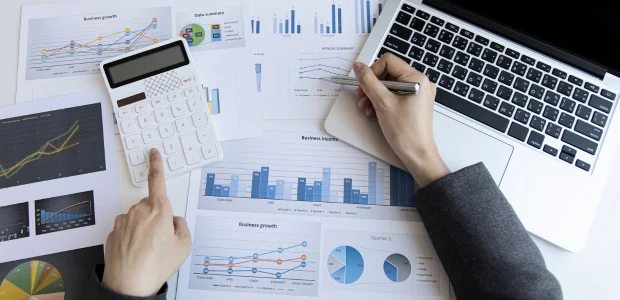 Symbolbild: Schreibtisch mit Taschenrechner, Laptop und wirtschaftlichen Auswertungen